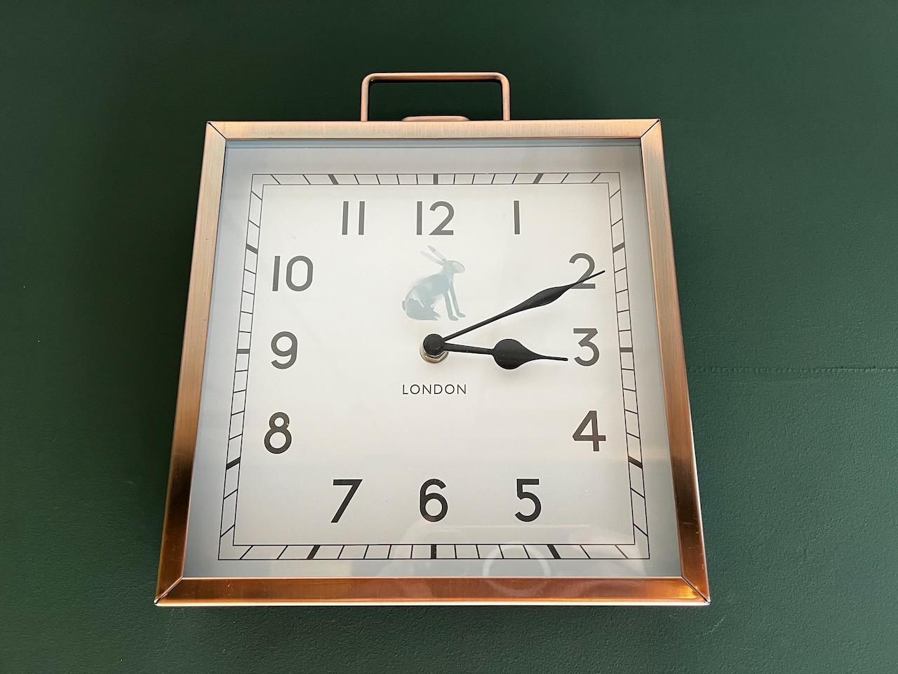 A copper-framed square clock on a dark green wall