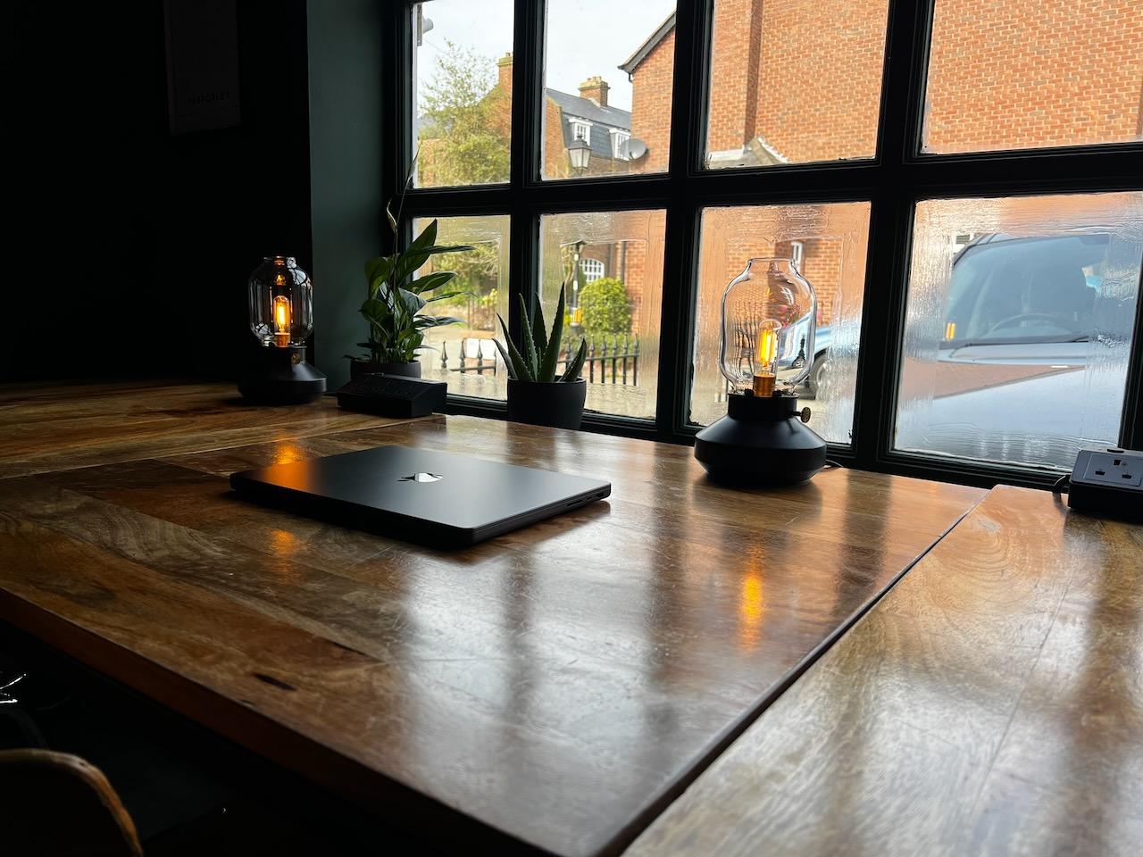 A closed laptop on a wooden desk by the window, lit by small glass table lamps