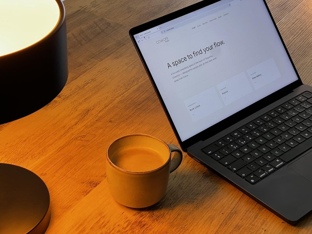 An open laptop showing the cowork.studio website, beside a cup of coffee and a lamp