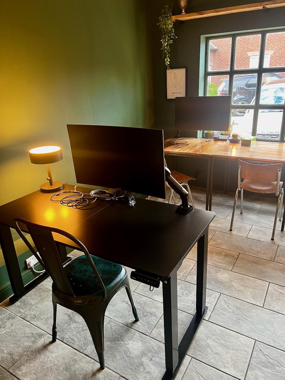 A sit-stand desk with a monitor on an arm and a desk lamp, with more desks by the window beyond