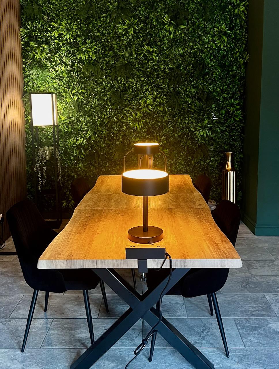 A wooden table with black chairs and a table lamp, in front of a wall of greenery