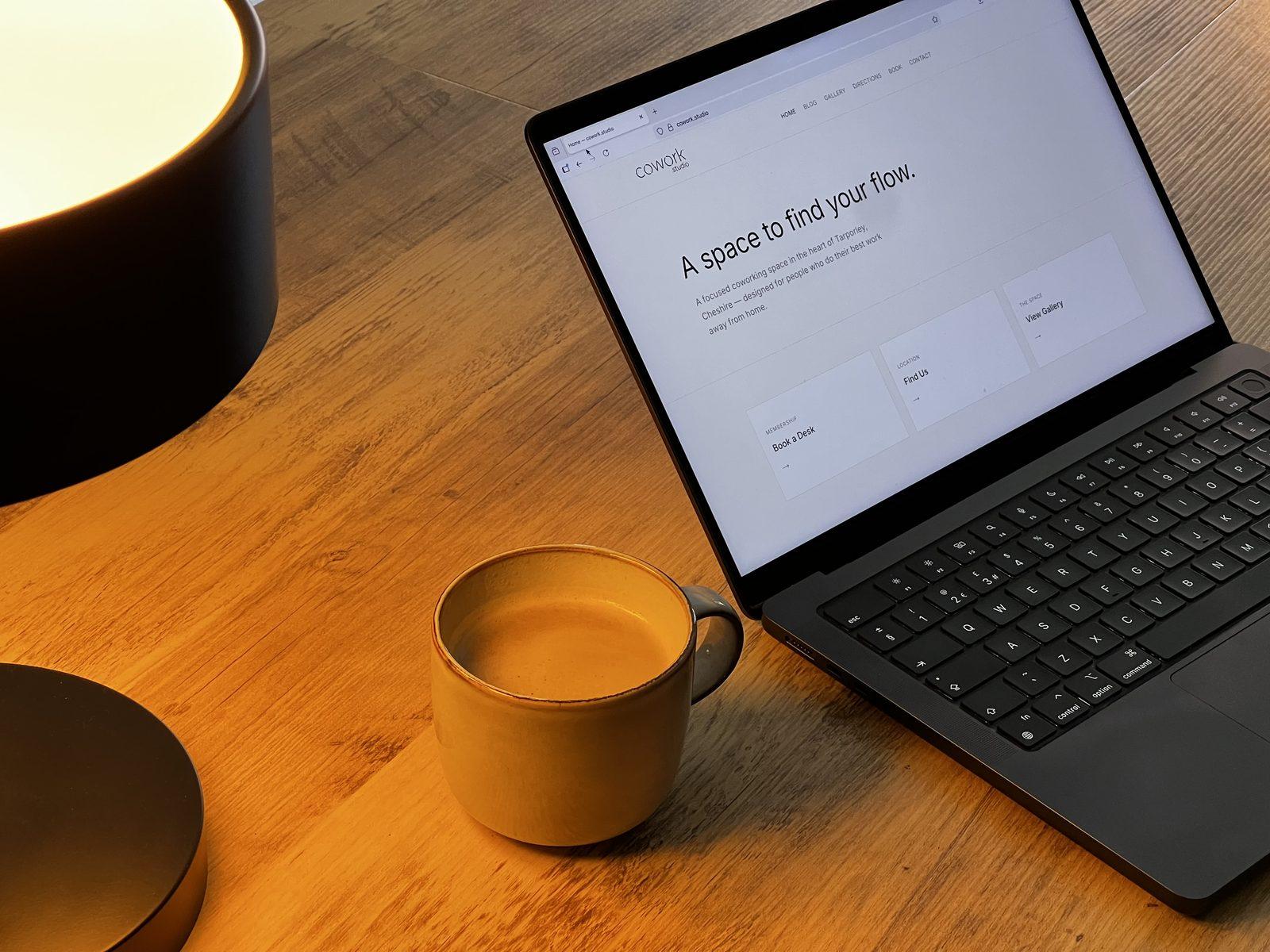An open laptop showing the cowork.studio website, beside a cup of coffee and a lamp
