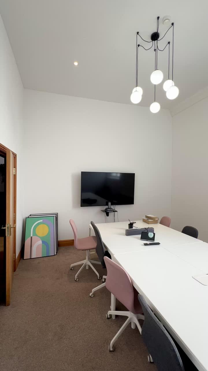 A long white meeting table with pink and grey chairs, facing a wall-mounted screen