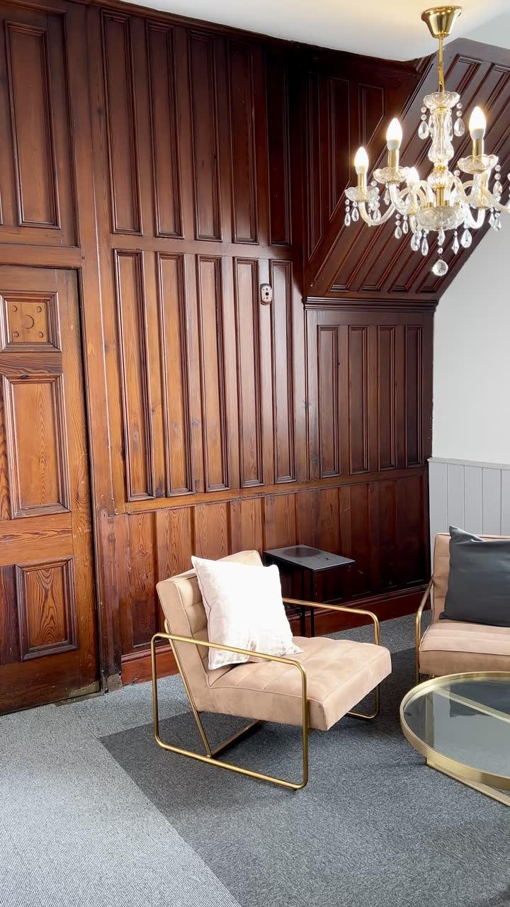 A pink armchair and a round glass table in a corner of dark timber panelling, under a cut-glass chandelier