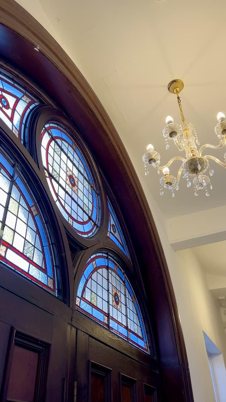 The arched stained-glass window above the chapel doors, lit blue and red, beside a cut-glass chandelier