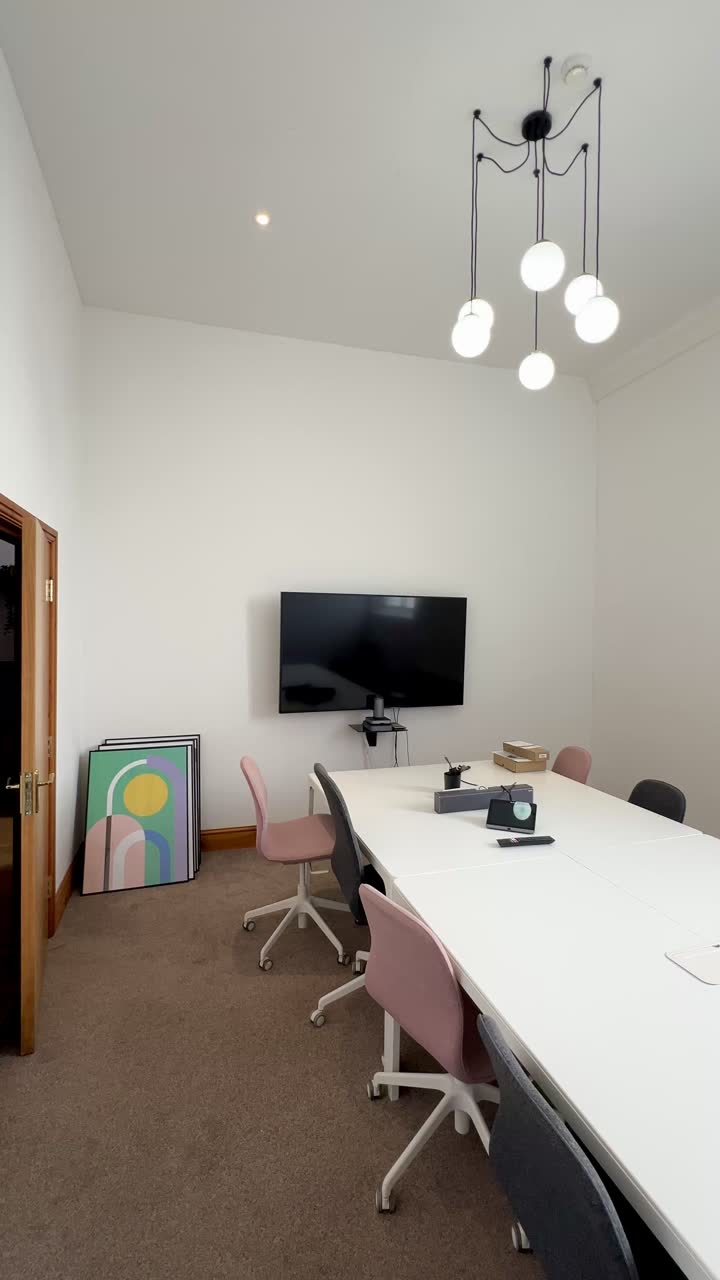 A long white meeting table with pink and grey chairs, facing a wall-mounted screen