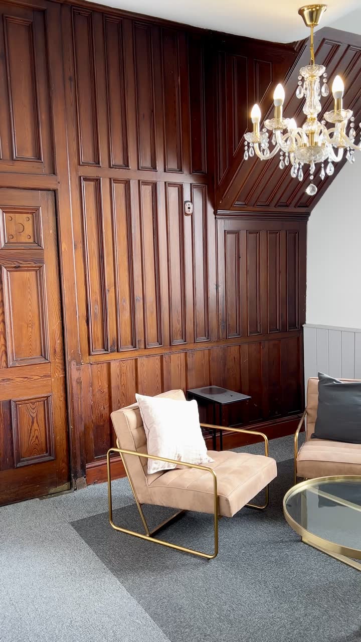 A pink armchair and a round glass table in a corner of dark timber panelling, under a cut-glass chandelier